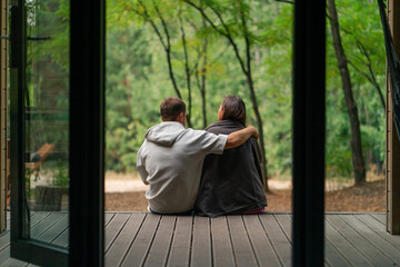 Man and woman in love sitting with their backs to the camera on a terrace overlooking  forest with plaids cuddling and talking - obrazy, fototapety, plakaty
