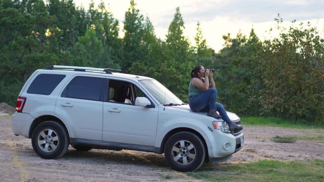 Woman with DSLR camera in her hands, sitting on the hood of her 4x4 SUV offroad car, taking photographs outdoors during sunset. - Long shot. Travel concept.