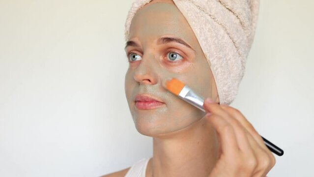 Attractive young woman applying clay facial mask as part of her skincare routine for healthy complexion wearing bath towel on head isolated over white background.