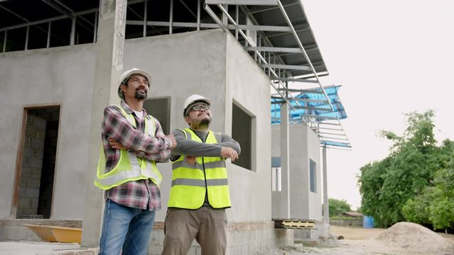 Two Asian construction engineers, standing, wearing hard hats protect their heads, when in construction zone for safety, engineers come stand supervise construction work that is large project.