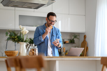 Happy young businessman eating cookie while checking messages over mobile phone at kitchen counter