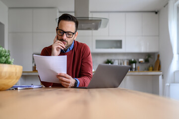 Serious handsome entrepreneur reading financial document while sitting with laptop at desk