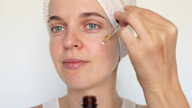 Face skin care. Cosmetology and beauty treatment. Woman wrapped in towel applying facial serum with dropper moisturizing skin domestic beauty routine isolated over white background.