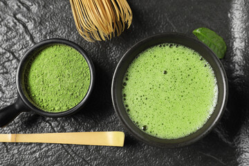 Cup of fresh matcha tea, bamboo whisk, spoon and green powder on black tray, flat lay