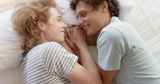 Happy diverse gay male couple lying on bed holding hands at home, slow motion