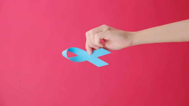 Vertical footage of female hand holding blue ribbon, isolated on red studio background wall with copy space. Healthcare, International men, Father and World cancer day concept. Symbol for support men