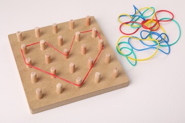 Wooden geoboard with heart made of rubber bands on white table. Educational toy for motor skills...