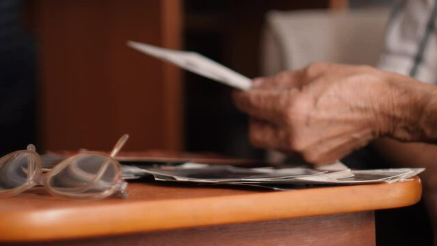an elderly man 80 years old looks at old family photos while sitting at the table.grandfather's hands holding old photos.selective focus.nostalgia concept, family history