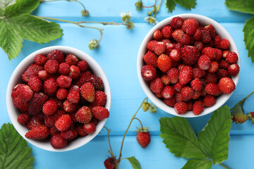 Fresh wild strawberries in bowls, flowers and leaves on light blue wooden table, flat lay