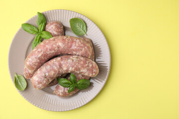 Raw homemade sausages and basil leaves on yellow background, top view. Space for text