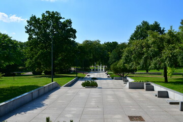 A close up on a well maintained park full of benches, fountains, shrubs, flower pots, forested areas and marble pavements seen on a sunny summer day near a small lake or river flowing through the area