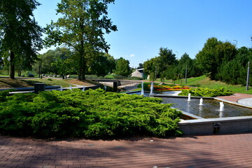 A close up on a well maintained park full of benches, fountains, shrubs, flower pots, forested areas and marble pavements seen on a sunny summer day near a small lake or river flowing through the area