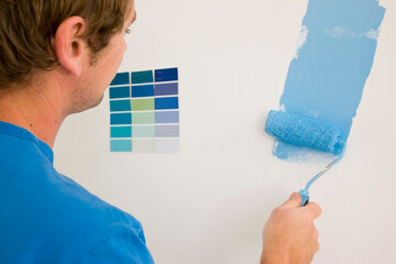 Back view of man holding paint roller and painting a white wall with blue paint
