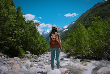 Young woman with backpack on her back stands on the rocks of mountain river and looks at the mountains. Active rest and relaxation