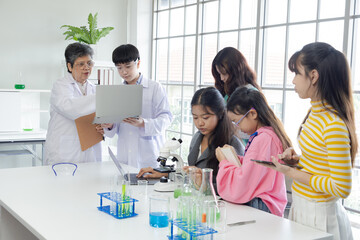 Group of Asian secondary students using technology laptop record the result in science laboratory class with senior 60s teacher wearing lab coat, healthy elderly woman working in modern private school