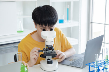 Cute schoolboy student sitting in science laboratory class looking to microscope analyzing using laptop technology record the experiment, new generation pupils in modern education learning lifestyle