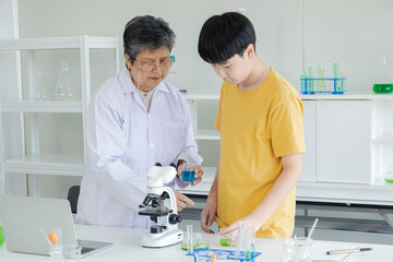 Asian cheerful schoolboy student using microscope standing with senior specialist teacher wearing lab coat in science laboratory, pupils learning by doing activities in class, elderly aging employment