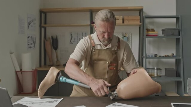 Medium shot of Caucasian adult man repairing knee of leg prosthesis, working alone in small prosthetics workshop