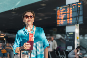 Tourist senior woman standing in airport hall in front of the timetable with a passport and plane...