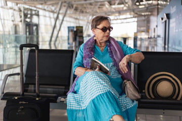 Tourist senior woman sitting on the bench in airport hall in watching her wristwatch waiting for her flight next to her suitcase