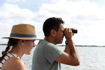 Obraz premium A man looks through a telescope at the horizon while a blurred woman wearing a hat looks in the same direction, with the ocean in the background.