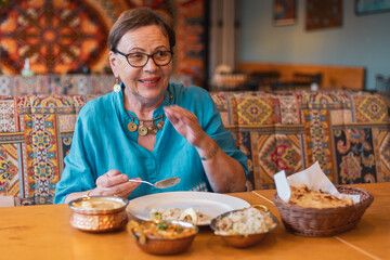 tourist senior woman laughing while eating delicious food in colorful indian restaurant