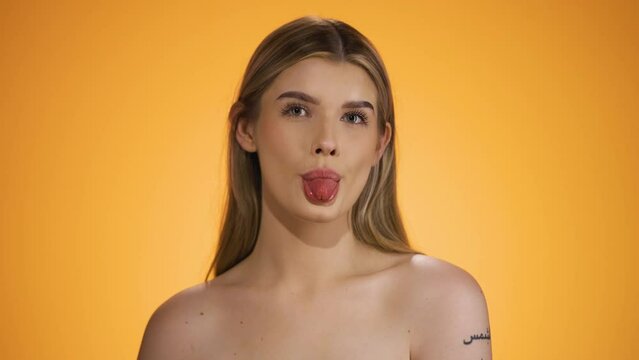 Medium shot of a young naked and pretty woman sticking out her tongue while looking ironically into the camera in front of yellow background in slow motion