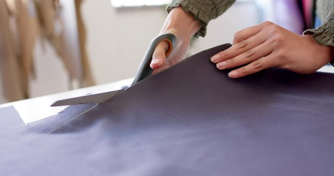 Hands of biracial female fashion designer cutting cloth with fabric shears on table, slow motion
