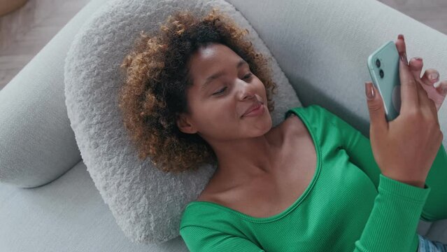 Young happy cute African American woman with smile falls on sofa and holds phone being carried away by correspondence in online messengers or taking selfie photo located in own apartment. Top view