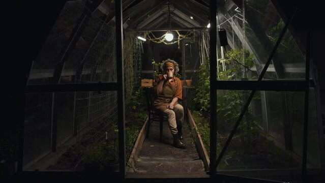 Wide shot of woman sitting on chair among tomato seedlings and drinking coffee at nighttime in hothouse