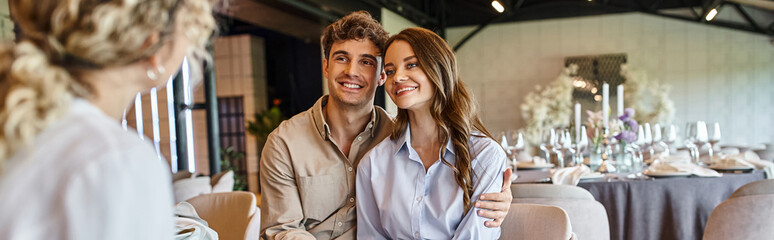 smiling couple looking at blurred event manager and sitting at festive table in wedding hall, banner