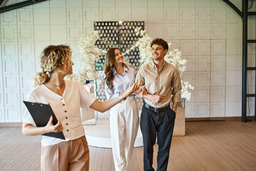 professional event manager with clipboard showing wedding venue with floral decor to happy couple