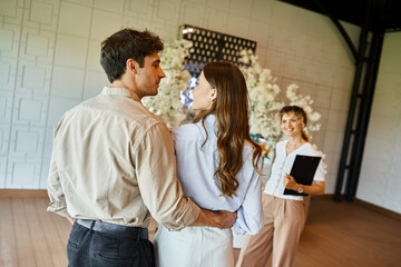 couple in love looking at each other near event coordinator in banquet hall with floral decoration
