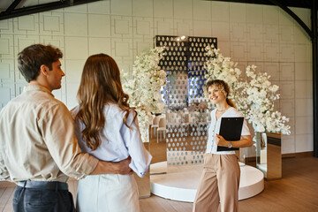 happy event manager with clipboard showing wedding venue with white floral composition to couple