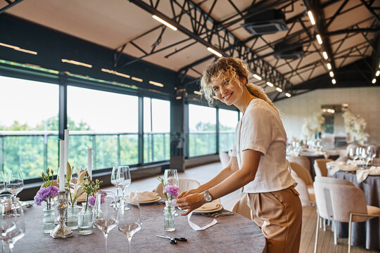 happy florist smiling at camera and decorating festive table with flowers for banquet in event hall