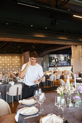 smiling event coordinator with clipboard talking on smartphone near festive tables in banquet hall