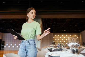 event coordinator with clipboard pointing with pen at table with festive setting in banquet hall