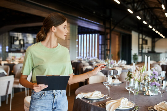 event manager holding clipboard and pointing with pen at table with festive setting in event hall