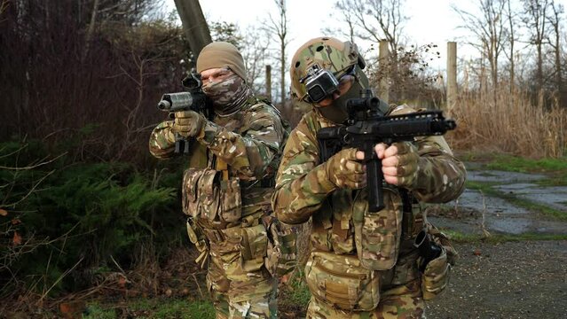 Two men in military camouflage uniforms with automatic rifles with optical sight aim at the background of an abandoned building. Airsoft soldier on battlefield.