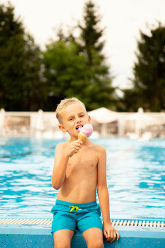 A seven-year-old boy of European appearance sits on the side of the pool and eats ice cream.