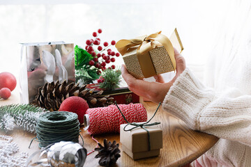 Woman in warm woolen sweater holding golden Christmas gift box in her hand on the background of dofferent bright festive decorations close up.