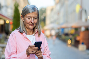 Senior smiling woman standing on city street and using mobile phone, typing messages, chatting, calling. Close-up photo.