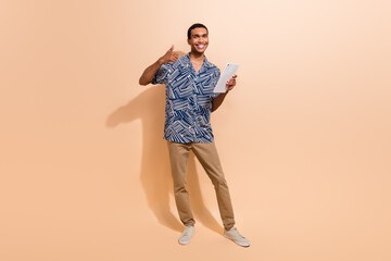 Fototapeta na wymiar Full length photo of satisfied man dressed stylish shirt brown pants showing thumb up holding tablet isolated on beige color background