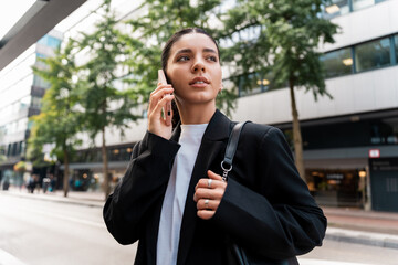 Wide portrait of siren businesswoman in black suit holding a phone to ear looking away in modern city street