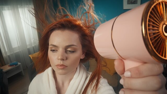 A close-up photo shows a young girl holding a hair dryer and drying her hair and styling it. Her gaze is directed downward, her hair is developing from the air flow. Sad expression on her face