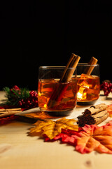 Vertical view of two glasses of whiskey with ice cubes and cinnamon sticks on a wooden table, with mapple leaves and black background