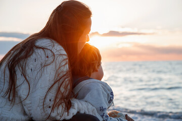 Mom and son are sitting on the seashore. The mother hugs the child. Happy childhood. Sunset in the...