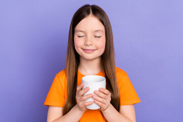 Photo of little schoolgirl closed eyes relax enjoy drinking sweet tea cup good mood wear orange t shirt isolated on violet color background