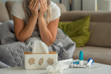 Medicines for colds and paper napkins are on the table, a woman with signs of flu and colds is sitting in the background and blowing her nose.