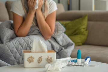 Medicines for colds and paper napkins are on the table, a woman with signs of flu and colds is sitting in the background and blowing her nose.